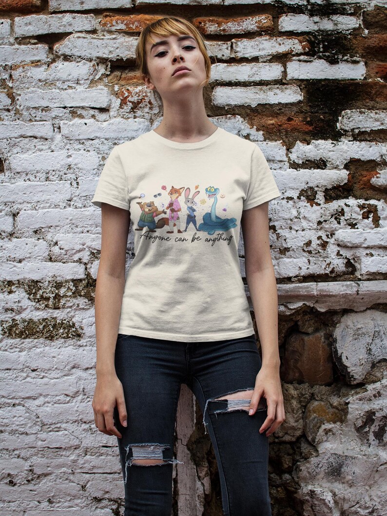 Pu&ograve; includere: Una t-shirt a maniche corte color crema con una stampa grafica di quattro personaggi animali animati e il testo "Anyone can be anything" scritto in corsivo sotto i personaggi. La maglietta ha un girocollo e un orlo dritto.