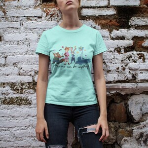 Pu&ograve; includere: Una t-shirt a maniche corte color verde menta chiaro con una stampa grafica di quattro personaggi animati e il testo "Anyone can be anything" in corsivo. La maglietta ha un girocollo e un orlo dritto, indossata con jeans blu scuro strappati.