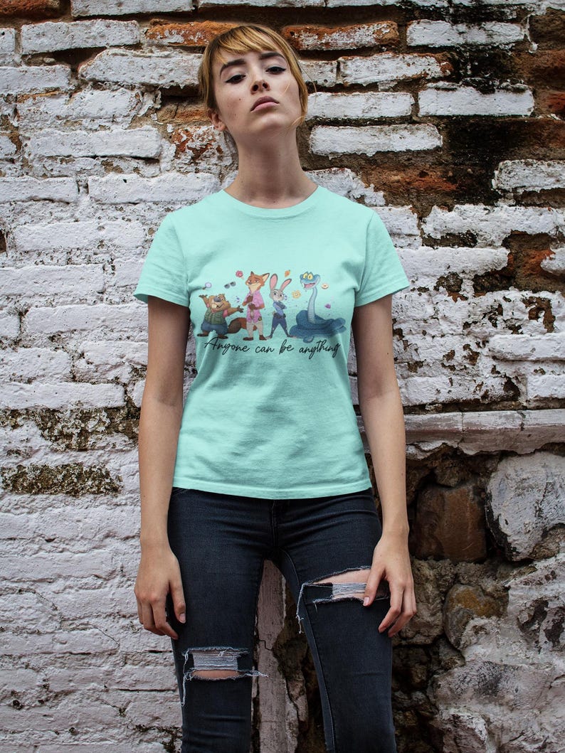 Pu&ograve; includere: Una t-shirt a maniche corte color verde menta chiaro con una stampa grafica di quattro personaggi animati e il testo "Anyone can be anything" in corsivo. La maglietta ha un girocollo e un orlo dritto, indossata con jeans blu scuro strappati.