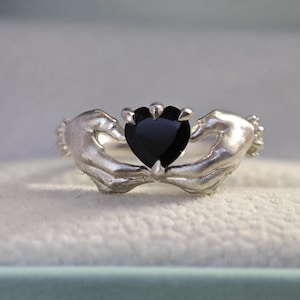 Può includere: Un anello Claddagh argentato con una pietra preziosa nera a forma di cuore. Il cuore è tenuto da due mani, un design tradizionale irlandese che simboleggia amore, lealtà e amicizia. L'anello è posto su uno sfondo morbido e neutro.