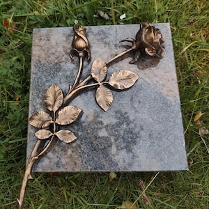 Op de afbeelding: Een bronzen metalen rozenbeeld met een knop en een volledig bloeiende bloem, samen met bladeren, is bevestigd op een vierkante, grijze marmeren basis. Het beeld staat tegen een achtergrond van groen gras.