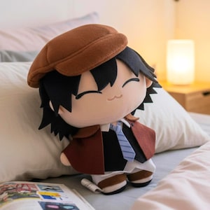 Puede incluir: Muñeco de peluche de un personaje de anime con sombrero marrón, capa roja oscura y corbata azul. El muñeco tiene pelo negro y una cara sonriente. El muñeco está sentado en una cama con una almohada blanca y un libro.