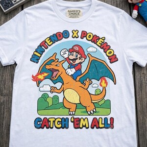 May include: A white short-sleeve t-shirt featuring a graphic of Mario riding Charizard. The text reads NINTENDO X POKÉMON and CATCH 'EM ALL! The design includes a blue sky, green hills, and clouds. The shirt has a Gamer's Choice label at the neck.