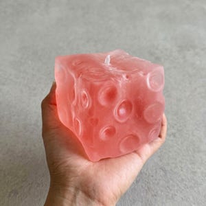 Pode incluir: Uma vela em forma de cubo na cor coral com uma superfície texturizada com reentrâncias circulares. A vela tem um pavio branco e é segurada na mão. Um item decorativo para casa.