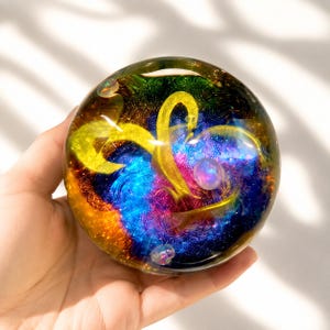 May include: A spherical glass paperweight featuring a swirling nebula-like design with vibrant streaks of yellow, deep blue, and magenta. The clear glass surface contains embedded glitter and small iridescent bubbles, creating a multi-dimensional effect.