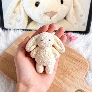 May include: A small, cream-colored crocheted bunny plush toy with long, floppy ears, a tiny pink nose, and two small dark eyes. The texture of the yarn is soft and fuzzy, and the bunny is held in a palm, showcasing its compact size and simple design.