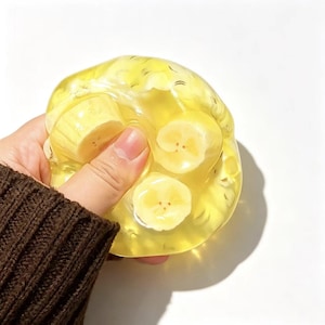 Op de afbeelding: Een doorschijnend geel, geleiachtig, rond sensorisch speelgoed met meerdere ingebedde plakjes banaan. Het materiaal is buigzaam en zacht, met een heldere, gelei-achtige textuur waardoor de gele en witte bananenstukjes duidelijk zichtbaar zijn.