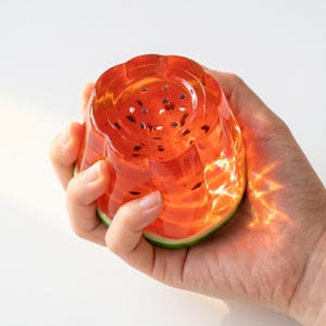 May include: A translucent, rounded object shaped like a watermelon slice, featuring a vibrant red interior with small black seed-like details and a thin green rind at the base. The material is glossy and catches light, creating bright reflections.