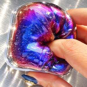 May include: A clear, round container filled with a thick, viscous substance featuring a vibrant galaxy-inspired colour palette of deep purple, royal blue, and warm orange hues, embedded with fine silver glitter particles throughout the texture.