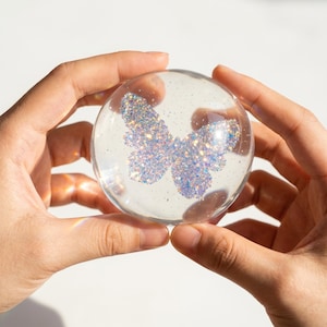 Puede incluir: Una esfera de vidrio transparente con un diseño de mariposa de purpurina en su interior. La mariposa está hecha de purpurina iridiscente, que refleja tonos azules, morados y plateados. La esfera se sostiene con dos manos.