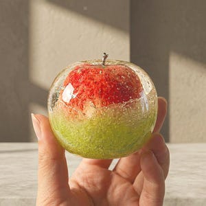 Pode incluir: Um peso de papel esférico transparente com um design de maçã vermelha e verde. A maçã é encapsulada em resina transparente, com a metade superior vermelha e a metade inferior verde. Uma pequena haste prateada é visível.