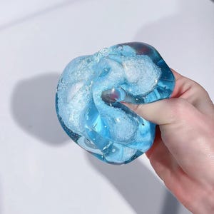 Könnte beinhalten: Ein durchscheinendes blaues Schleimspielzeug mit weißen Wirbeln und Glitzer wird in einer Hand gehalten. Das Spielzeug hat eine leuchtend blaue Farbe und scheint weich und biegsam zu sein. Der Hintergrund ist eine schlichte weiße Oberfläche.