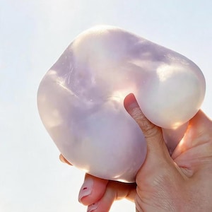 May include: A translucent, pale lavender squishy toy with a smooth, gel-like texture. The material is pliable and semi-transparent, allowing light to pass through its rounded, irregular shape while being squeezed to show its flexible consistency.