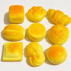 May include: A collection of nine yellow foam squishy toys shaped like various baked goods, including a square loaf, a paw print, a croissant, an oval roll, a round bun, a textured square, a braided pastry, and a smooth round bread roll.