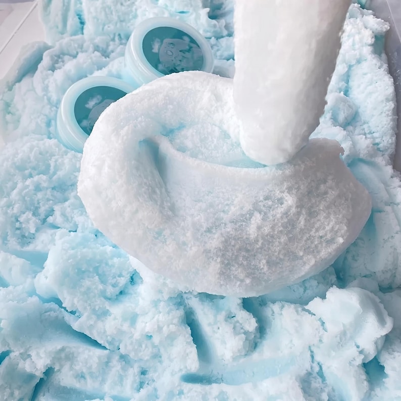 May include: A close-up of blue and white fluffy slime with two small containers. The slime has a textured, cloud-like appearance. The containers have blue lids with a white image.