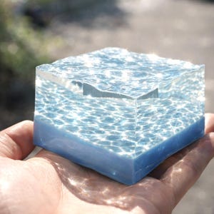 May include: A clear, cube-shaped resin art piece with a blue base and water-like patterns. The top surface mimics rippling water, with light reflecting off the surface. The artwork is held in a hand, showcasing the miniature sculpture.