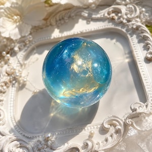以下が含まれることがあります： 半透明の青いベースに、渦巻く金色のメタリックなアクセントとグリッターの粒子が埋め込まれた、滑らかな球体の樹脂製オーブ。この球体は、明るい青から暗い青へと変化する、海のような色のグラデーションを示しています。