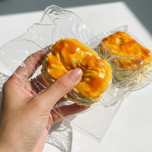 May include: Two round, textured, light-colored baked goods topped with a glossy, vibrant orange glaze, each individually sealed in a clear, crinkled plastic bag. The surface of the baked items appears uneven and porous under the plastic.