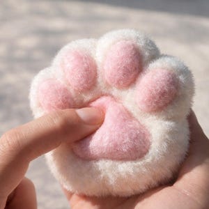 Könnte beinhalten: Nahaufnahme einer weichen, weißen Katzenpfote mit rosa Ballen. Ein Finger drückt sanft auf die Mitte der Pfote. Das Bild zeigt die flauschige Textur und das detaillierte Design. Es handelt sich wahrscheinlich um einen Neuheits- oder Komfortartikel.