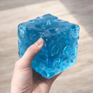 May include: A translucent, bright blue cube with a textured surface featuring numerous circular indentations. The object has a glossy, glass-like appearance and a distinct geometric shape, reflecting light across its uneven, dimpled exterior.