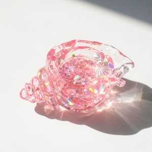 May include: A pink, seashell-shaped resin dish filled with pink glitter, iridescent flakes, and miniature seashell and star embellishments. The dish is translucent, allowing light to pass through, and is set against a white background.