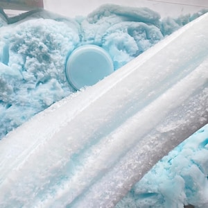 May include: A close-up of a container filled with light blue slime. The slime has a fluffy, textured appearance, with a smooth, light blue lid in the center. A white, textured substance is layered diagonally across the image.