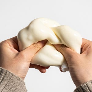 Peut inclure: Un objet souple et blanc cassé est tenu dans deux mains. L'objet est tordu et noué, avec une surface lisse et brillante. Les mains portent un pull beige. Le fond est blanc uni.