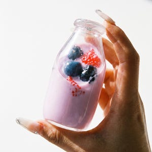 May include: A small, clear glass bottle filled with a light purple yogurt or smoothie base. The drink is topped with several whole dark blue blueberries and a cluster of bright red raspberries, creating a vibrant contrast against the pale purple liquid.