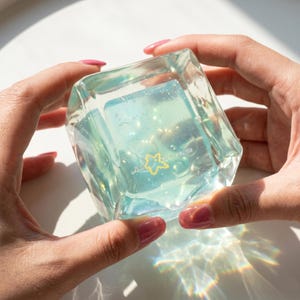 May include: A clear, cube-shaped object with a light blue interior and a yellow star design. The object is held between two hands, with the light reflecting off the surface. The object appears to be made of glass or a similar transparent material.