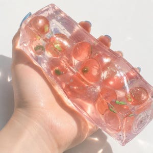 Può includere: Un blocco rettangolare in resina rosa trasparente contenente diversi oggetti sferici incorporati che ricordano piccoli frutti con steli verdi. Il materiale trasparente è infuso con minuscoli glitter argentati a forma di stella.
