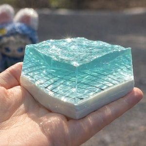 May include: A square, translucent sculpture with a blue-green, water-like top layer and a grid pattern beneath. The base is white. The object is held in a hand, with a blurred background.