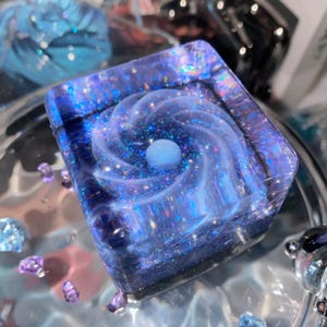 Pode incluir: Um peso de papel quadrado em resina com um padrão de redemoinho inspirado numa galáxia em tons de azul profundo e roxo. O material translúcido está incrustado com purpurina holográfica fina e uma esfera azul clara situa-se no centro.