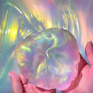Peut inclure: Une pâte visqueuse translucide et irisée avec une texture gélatineuse brillante qui reflète des couleurs arc-en-ciel vibrantes, notamment le jaune, le rose et le bleu. Le matériau semble malléable et lisse, captant la lumière.