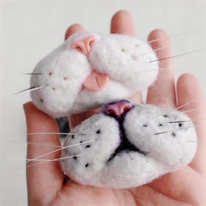 Puede incluir: Dos caras de gato de fieltro blanco con narices rosas y bigotes. Las caras están detalladas con pequeños puntos negros y se sostienen en una mano. Las caras de gato son un artículo artesanal.
