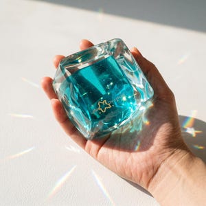 Könnte beinhalten: Ein klarer, facettierter Glasbehälter mit türkisem Flüssigkeitsinneren. Der Behälter hat ein sternförmiges goldenes Emblem mit Text. Das Objekt wird in einer Hand vor weißem Hintergrund gehalten, wobei sich das Licht durch das Glas bricht.