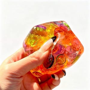 May include: A clear, multi-faceted resin cube containing numerous small, colourful, translucent plastic buttons in shades of orange, yellow, red, and purple. The object has a smooth, glossy finish and a geometric shape with rounded edges.