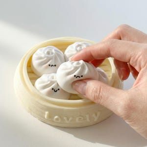 May include: A cream-coloured bamboo steamer containing several white, dumpling-shaped objects, each with a small, black, cartoon-style face. One is being held by a hand. The word "lovely" is printed on the steamer.