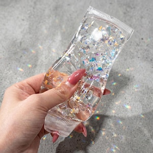 Pode incluir: Um tubo flexível de plástico transparente preenchido com pequenas formas geométricas facetadas e iridescentes que refletem a luz num espectro de arco-íris. O recipiente apresenta um selo estriado na parte superior e inferior.
