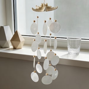 Pode incluir: Um carrilhão decorativo com um topo em forma de estrela-do-mar e cordas de conchas brancas em forma oval. O carrilhão está pendurado em frente a uma janela, com dois vasos geométricos e um copo na janela.