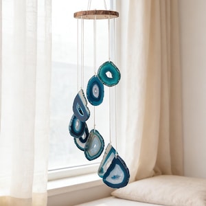 May include: A hanging wind chime featuring slices of blue and white agate stones suspended from a wooden ring. The agate slices vary in shape and size, creating a decorative and natural aesthetic. The chime hangs in front of a window.