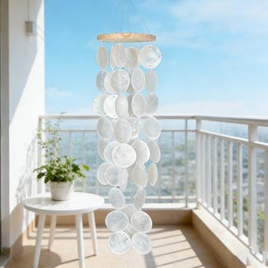 Puede incluir: Un carillón de viento decorativo hecho de conchas de capiz blancas, suspendido de un anillo de madera. El carillón está compuesto por numerosas conchas redondas y planas. El fondo muestra un balcón con una barandilla blanca y un cielo azul claro.
