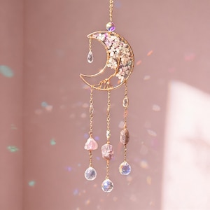 Puede incluir: Un atrapasoles dorado en forma de media luna con un diseño floral y cristales colgantes. El atrapasoles tiene una delicada cadena para colgar y presenta varias formas y colores de cristal, incluyendo transparente, rosa y marrón.