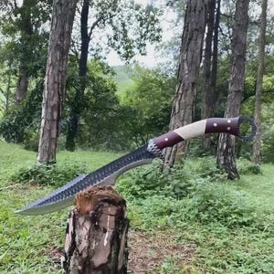Può includere: Un coltello a lama curva di grandi dimensioni con bordo seghettato e manico in legno. Il manico è una combinazione di legno chiaro e scuro con un anello all'estremità. Il coltello è esposto su un ceppo d'albero in un ambiente forestale.