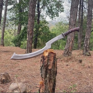 May include: A curved metal blade with a sharp, pointed tip and a dark brown handle featuring silver rivets. The blade has a distinct, sweeping design with a metallic finish, and the handle is secured with multiple metal pins along its length.