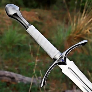 Può includere: Una spada con pomolo in metallo argentato lucido, impugnatura avvolta in bianco e guardia con un piccolo inserto circolare blu al centro. La lama è realizzata in metallo argentato riflettente con una scanalatura centrale lungo la lunghezza.