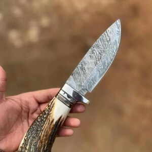 Peut inclure: Un couteau à lame fixe doté d'une lame en acier damas argenté avec un motif ondulé distinct. Le manche est fabriqué à partir d'un matériau en bois de cerf texturé, brun et crème, séparé de la lame par une mitre argentée polie.