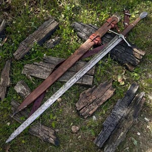 May include: A long sword with an etched metal blade featuring intricate patterns, a silver crossguard, and a brown wooden handle. Beside the sword lies a matching brown leather scabbard with leather straps and metal buckles for secure attachment.