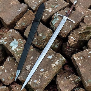 May include: A long, double-edged steel sword with a pointed tip and a central fuller. The hilt features a dark brown wooden grip with silver-toned metal accents and a crossguard. Beside it lies a black scabbard wrapped in black leather straps.