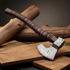 May include: A handcrafted axe with a patterned steel blade and a dark brown leather-wrapped handle. The axe head features a unique swirling design. The handle is wrapped with dark brown leather strips. The axe is displayed on wooden planks.