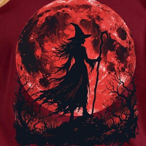 May include: A maroon short-sleeve t-shirt featuring a graphic print of a witch in a pointed hat holding a staff, standing before a large, textured red moon. The silhouette of the witch is black, set against a dark, barren landscape of trees.